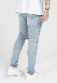 Jeans de mezclilla azul claro con detalles desgastados, que presentan salpicaduras de pintura en rojo y blanco, corte slim, con puños doblados y bolsillos traseros.