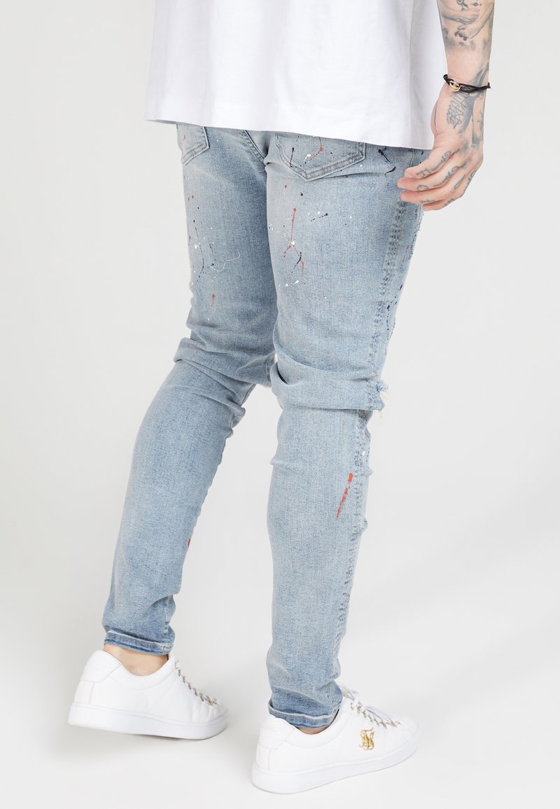 Jeans de mezclilla azul claro con detalles desgastados, que presentan salpicaduras de pintura en rojo y blanco, corte slim, con puños doblados y bolsillos traseros.