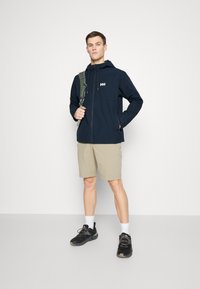Chaqueta con cremallera azul marino y capucha, con logo en el pecho, combinada con pantalones cortos beige y zapatillas deportivas negras. El modelo sostiene la correa de una bolsa verde.
