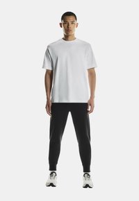 Witte katoenen T-shirt met korte mouwen, gecombineerd met zwarte getailleerde sweatpants. Model draagt witte en zwarte sport schoenen.