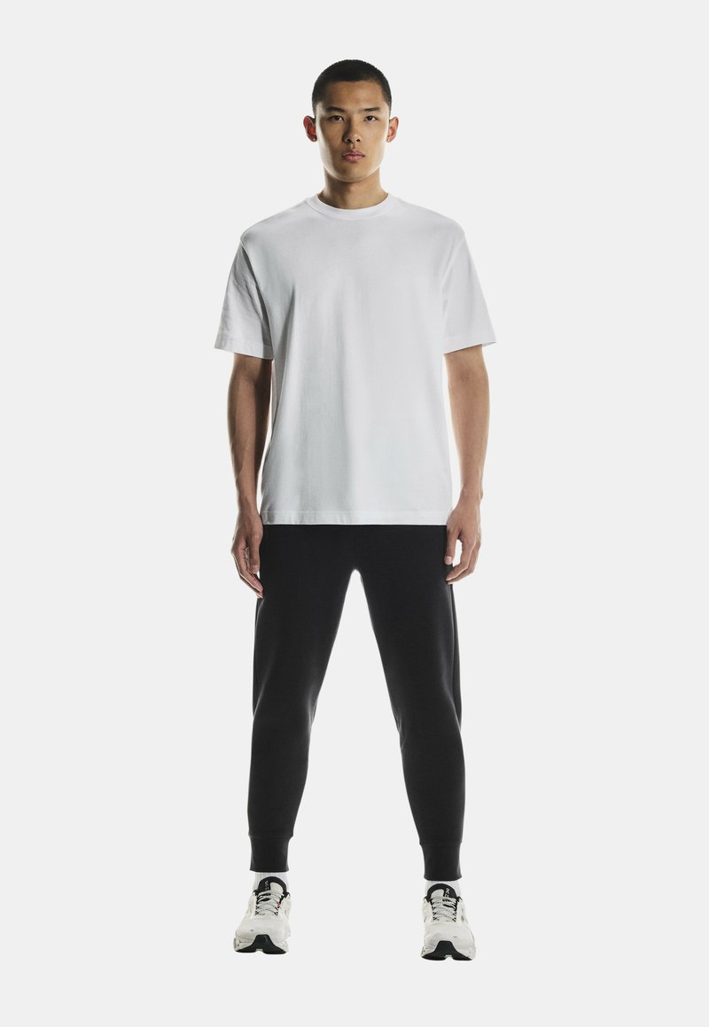T-shirt en coton blanc à manches courtes, associé à un pantalon de jogging noir fuselé. Modèle portant des chaussures de sport blanches et noires.