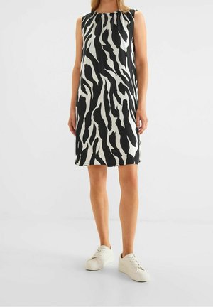 Kobieta w czarno-białej sukience w wzór zebry, bez rękawów, sięgającej kolan, nosząca białe trampki, na jednolitym tle.