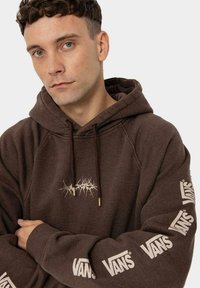 Brązowa bluza z kapturem ze ściągaczami; na piersi widoczny beżowy, wyraźny logotyp, a na rękawach powtarzające się oznaczenia marki; miękka bawełniana faktura.