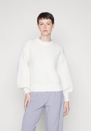 Pull blanc en tricot avec des côtes diagonales, des manches ballon et un ourlet côtelé. Associé à un pantalon gris clair ample.