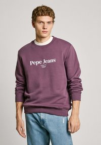 Paarse sweatshirt gemaakt van zachte katoen met een ronde hals en geribde manchetten. Heeft een wit logo en tekst op de voorkant. Draagt met blauwe jeans.