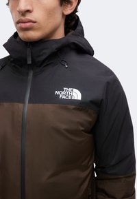 Άνδρας που φοράει μαύρο και καφέ μπουφάν North Face με ψηλό γιακά και ορατό λογότυπο στο στήθος μπροστά σε απλό φόντο.