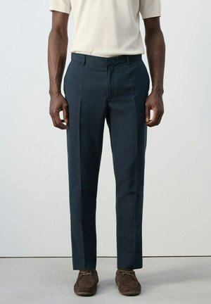 Hombre con pantalones ajustados de color azul marino oscuro, zapatos de ante marrones y una camisa ligera de manga corta, de pie frente a un fondo liso.