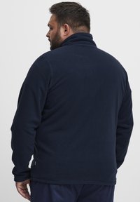 Suéter de forro polar azul marino con un cuello alto, tejido texturizado, mangas largas y una abertura lateral para facilitar el movimiento. Se muestra la vista posterior.