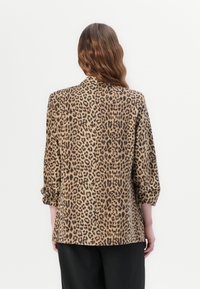 Žena s dlouhými vlnitými vlasy nosí hnědo-černou blůzu s leopardím potiskem a černé kalhoty, otočená zády k jednobarevnému bílému pozadí.
