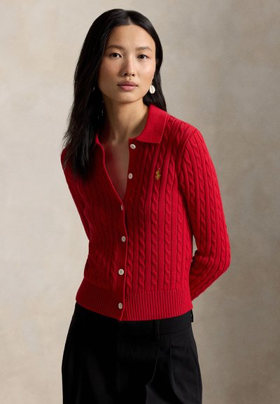 Woman with long black hair wears a red cable-knit cardigan with white buttons and black pants, standing against a neutral background.