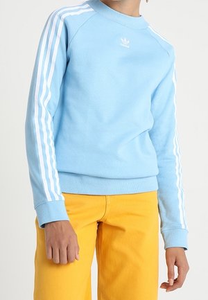 Jasnoniebieska bluza Adidas z białymi paskami na rękawach, noszona z musztardowożółtymi spodniami, pokazana od ramion do bioder.