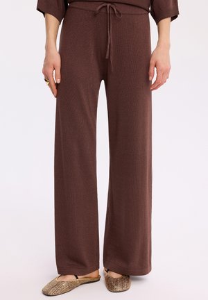 Brązowe spodnie o szerokich nogawkach z talią na sznurek, noszone z plecionymi płaskimi butami typu slip-on na osobie stojącej na białym tle.