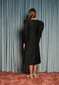 Svart teksturert midikjole med pufferm, med innsvingt midje og glatt stoff. Brukt med hvite blokkhælte sko foran et drapert bakteppe.