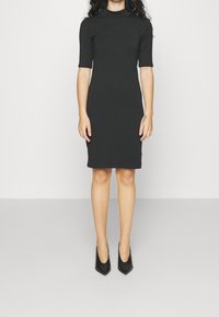 Svart figursydd midiklänning med halvärmar och hög krage, tillverkad av ett slätt tyg. Matchad med spetsiga svarta klackar.