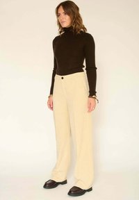 Pull en maille côtelée marron, pantalon large en velours côtelé crème, chaussures noires à texture, et accessoires en or aux poignets.