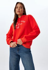 Czerwona bluza z kapturem z białym napisem "NEW YORK BRONX 86". Posiada regulowany kaptur i prążkowane mankiety, noszona z niebieskimi dżinsami.