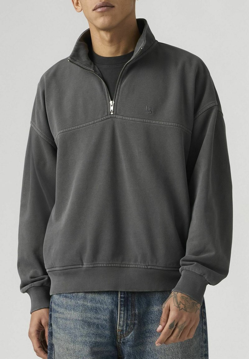 Homme portant un pull à demi-fermeture éclair gris foncé avec un petit logo "LS" et un jean bleu clair, montrant sa main droite tatouée.