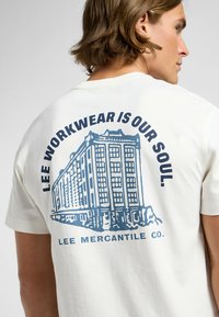 Hvid bomulds t-shirt med et blåt grafisk motiv af en bygning og teksten "LEE WORKWEAR ER VORES SJÆL. LEE MERCANTILE CO." på ryggen.