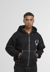 Černá mikina na zip s bílými švy, předními kapsami a grafickým logem na levé hrudi. Materiál vypadá měkce a texturovaně.