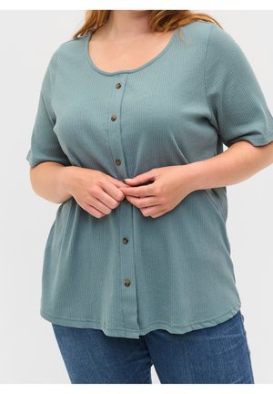Frau trägt ein blau-grünes, geripptes, kurzärmeliges Oberteil mit Knöpfen und blaue Jeans, richtet die Knöpfe vorne am Oberteil.