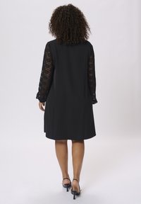 Abito nero con maniche lunghe in pizzo, lunghezza al ginocchio, texture liscia, vestibilità rilassata e tasche laterali discrete. Indossato con tacchi a strappo neri.