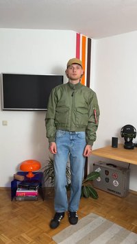 Jachetă bomber verde cu două buzunare frontale, închidere cu fermoar și manșete elastice, purtată peste blugi albaștri. Sneakers negri și o șapcă bej completează ținuta.