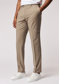 Beige maßgeschneiderte Hosen aus glattem Stoff, gerader Schnitt, mit frontalen Taschen und subtiler Textur, kombiniert mit weißen Sneakers.