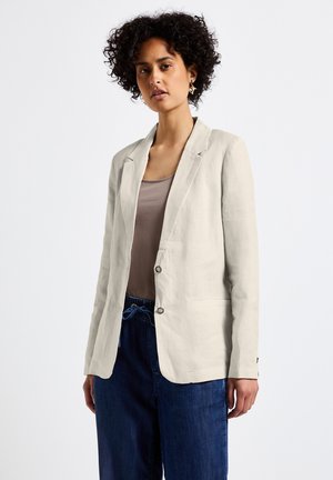 Blazer en lin beige clair avec une coupe ajustée, fermeture à un bouton, revers crantés, poches latérales et une texture lisse. Associé à un jean foncé.