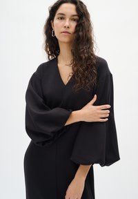 Fekete ruha V nyakkivágással és dús ujjal. Puha anyagból, sima textúrával készült. Egyszerű, elegáns kialakítással rendelkezik.