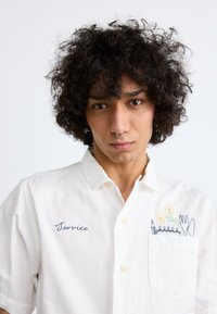 White button-up shirt with short sleeves, featuring blue embroidered text and floral design on the pocket. Soft cotton fabric.