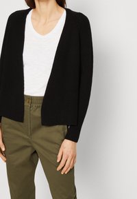 Svart stickad kofta med öppen framsida, ribbad textur och långa ärmar, bärs över en vit t-shirt med v-hals och olivgröna byxor.