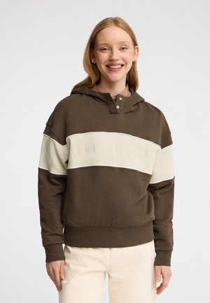 Joven sonriente con una sudadera con capucha marrón y beige con texto "POLO CLUB" y pantalones de color claro sobre un fondo liso.