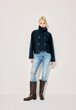 Žena oblečená v čiernej bunde s vysokým golierom zapínateľnej na gombíky, svetlé roztrhané úzke džínsy a vysoké tmavohnedé čižmy, stojaca opretá o obyčajnú stenu.