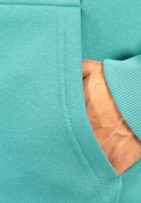 Turquoise sweatshirtmateriaal met een voorknoopzak. Kenmerkt zich door ribgebreide manchetten en een gladde textuur, benadrukt door zichtbare stikseldetails.