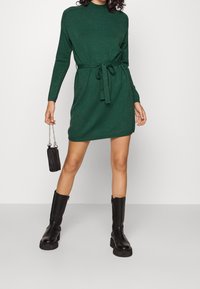 Robe en maille verte à manches longues et col montant, cintrée à la taille avec un lien assorti. Associée à des bottes noires montantes et un petit sac.