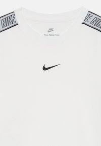 Balta Nike marškinėliai su juodu swoosh logotipu, esantis centre po apykakle, ir prekės ženklu juodai baltais juostelėmis ant pečių.