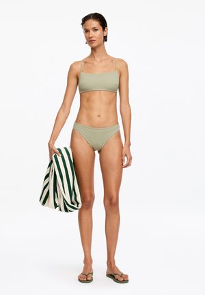 Frau trägt einen hellolivfarbenen Bikini und Flip-Flops, hält ein weißes Handtuch mit dunkelgrünen Streifen, steht vor einem schlichten weißen Hintergrund.