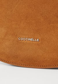 Materiale in suede marrone con una texture morbida. Presenta il logo "Coccinelle" in argento in rilievo. Cuciture lisce lungo il bordo.