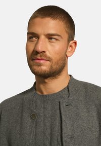 Šedý pletený cardigan s kulatým výstřihem, který má dekorativní detaily knoflíků a jemný vzor kabelkového pletiva. Měkká, texturovaná tkanina.