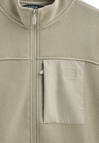 Giacca zip-up grigio chiaro in materiale a maglia testurizzata, colletto rialzato e una tasca a toppa in tessuto liscio a contrasto con logo.