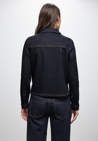 Giacca di jeans scura con un design aderente, caratterizzata da un colletto classico, maniche lunghe e cuciture in evidenza. La vista posteriore mostra due tasche sul retro.