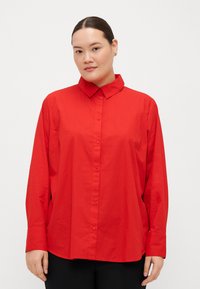 Camicia rossa con bottoni, colletto classico, maniche lunghe e vestibilità ampia. Tessuto liscio senza motivi o dettagli visibili.