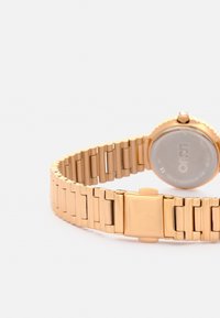 Reloj de pulsera de metal en tono dorado con una pulsera de eslabones y una caja redonda. Presenta un bisel liso y un cierre seguro. La parte trasera muestra los detalles de la marca.