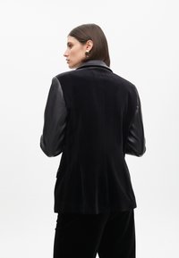 Blazer de terciopelo negro con mangas de cuero, corte entallado, solapa de muesca y textura suave; diseño minimalista, color sólido, sin patrones.