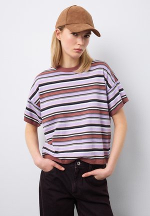 Joven con gorra marrón, camisa de manga corta a rayas en malva, púrpura y negro, con pantalones negros, de pie contra una pared lisa.