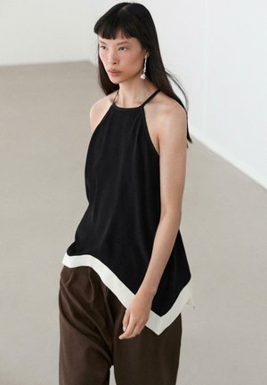 Frau mit langen schwarzen Haaren, die ein ärmelloses schwarzes Oberteil mit weißem Besatz und braune weite Hosen trägt, steht in einem minimalistischen Innenraum.