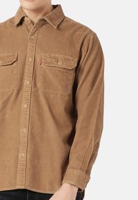 Brown corduroy shirt with two front pockets, button-down collar, and long sleeves. Features textured fabric with subtle vertical stripes.