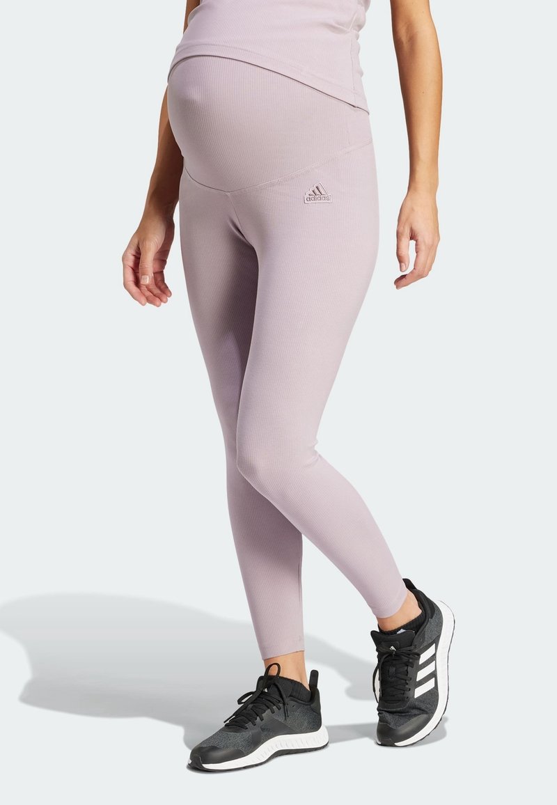 Gravid person klädd i ljuslila Adidas gravidleggings och svarta Adidas sneakers, stående med ena foten lätt upphöjd.