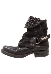 Bota de cuero negro con diseño holgado, adornada con tachuelas de metal en el puño, una correa con herrajes y una punta elegante curva.