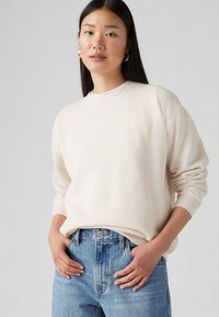 Felpa color crema con collo rotondo e polsini a coste, abbinata a jeans a vita alta azzurri chiari. Dettaglio del logo discreto sul petto.